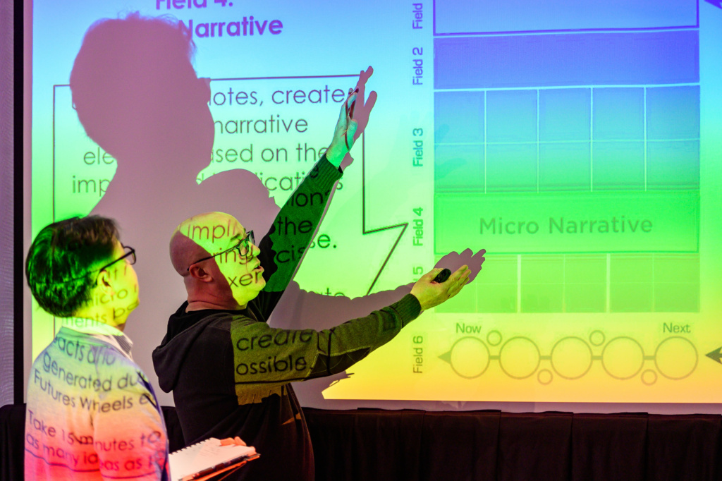 A colorful photo of Frank demonstrating the Scenario Fields diagram to a participant, the projection light covering both of them in colorful beams of light.